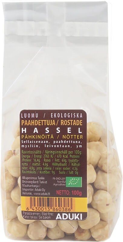 A clear plastic bag of Aduki organic roasted hazelnuts, rectangular in shape, with a matte finish label showing Finnish and Swedish text, nutritional information table, barcode, and 100g net weight. The label uses sans-serif font in black on a white background. No people or hands visible. Neutral, plain background in a close-up product shot.