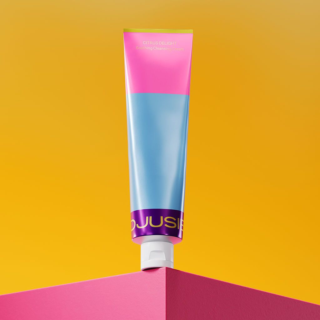 A colorful tube of cream featuring pink, blue, and metallic purple sections stands upright on a bright pink corner against a yellow background. The tube is made of glossy plastic, cylindrical with a flat flip-top cap at the base. Packaging displays segmented colors without gradients. No visible people or hands. Brand label text uses bold sans-serif font; only product name and logo are legible on the front.