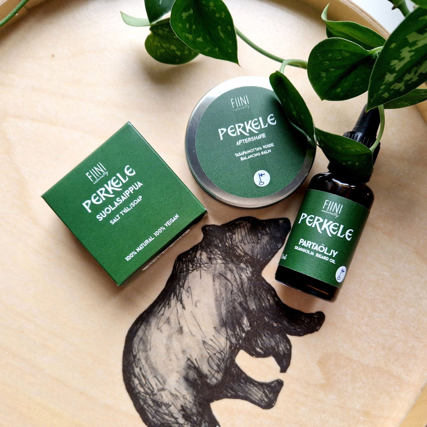 Three green-labeled Finnish skincare products are arranged on a wooden tray featuring an illustration of a bear, beside a lush green leafy plant. The selection includes natural soap, aftershave balm, and beard oil. Labels have simple sans-serif fonts and some Finnish text. Bottles and box shapes include cylindrical glass with matte finishes and a rectangular cardboard box. No people or hands present.
