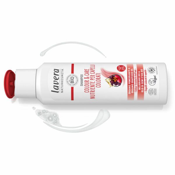 A white plastic bottle of Lavera Color & Care Shampoo with a red flip-top cap is shown upright. The bottle has a matte surface and a slightly curved rectangular shape. The front label displays pomegranate and quinoa illustrations and includes clear, sans-serif text for the product name. Swirls of white foam are beside the bottle on a plain, light background.