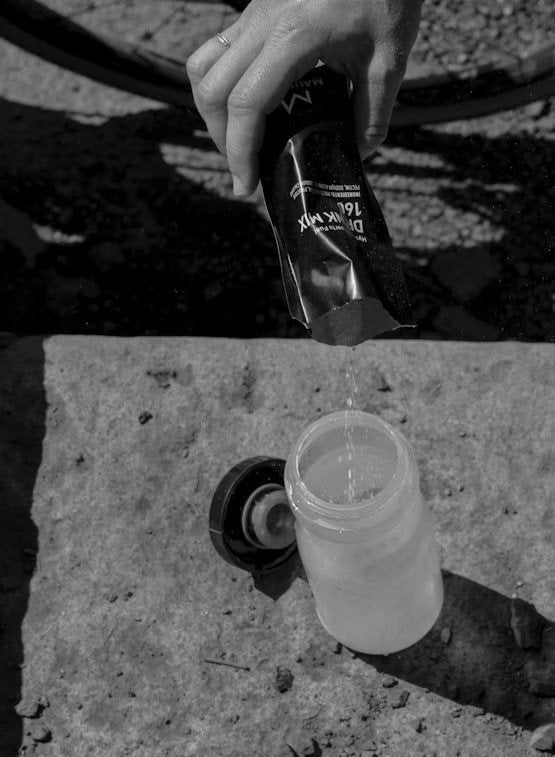 A hand pours powdered drink mix from a packet into a clear plastic water bottle with the white cap set aside. The packet is small, rectangular, and features printed text. The water bottle is cylindrical with smooth sides. All items are on an outdoor stone surface. No other objects or text are visible in the frame.