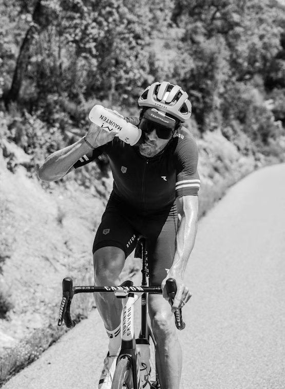 A cyclist wearing a helmet and sunglasses rides uphill on a road, drinking from a water bottle, with trees and bushes in the background. The black and white image shows one person in athletic clothing. Surface details of the bike are visible but not branded. There is no packaging or prominent text in the image. Outdoor setting, action shot, full body visible.