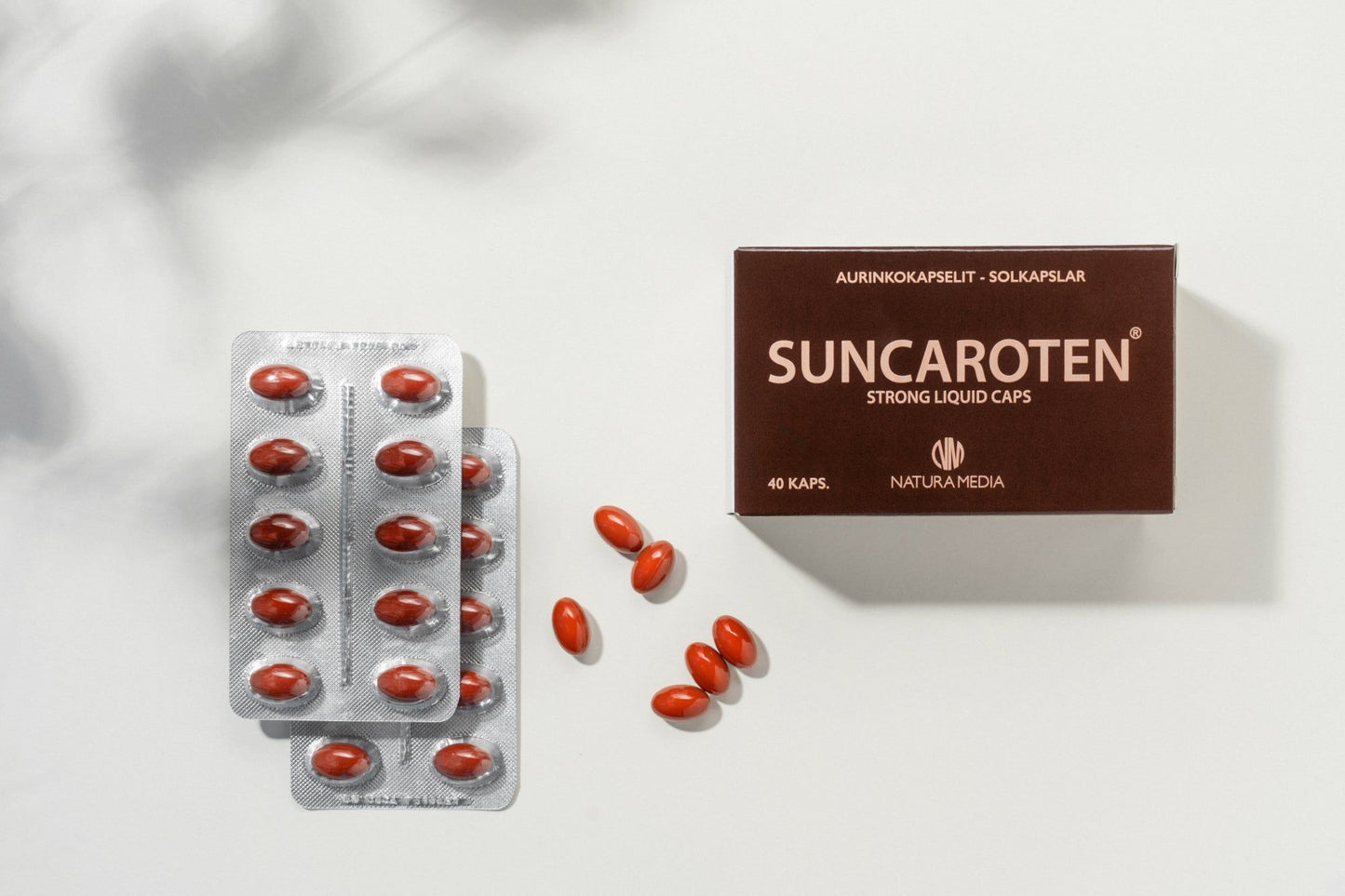 Natura Media SunCaroten Aurinkokapselit; lÀpipainopakkauksia ja irrallisia punaisia kapseleita ruskean Suncaroten-pakkauksen vieressÀ valkoisella alustalla; laatikko suorakulmainen mattapintainen, valkoinen lihavoitu groteskiteksti kertoo tuotteen sisÀltÀvÀn vahvoja karotenoidikapseleita, 40 kappaletta; ei ihmisiÀ tai kÀsiÀ; kuvattu ylhÀÀltÀ valkoisella taustalla.