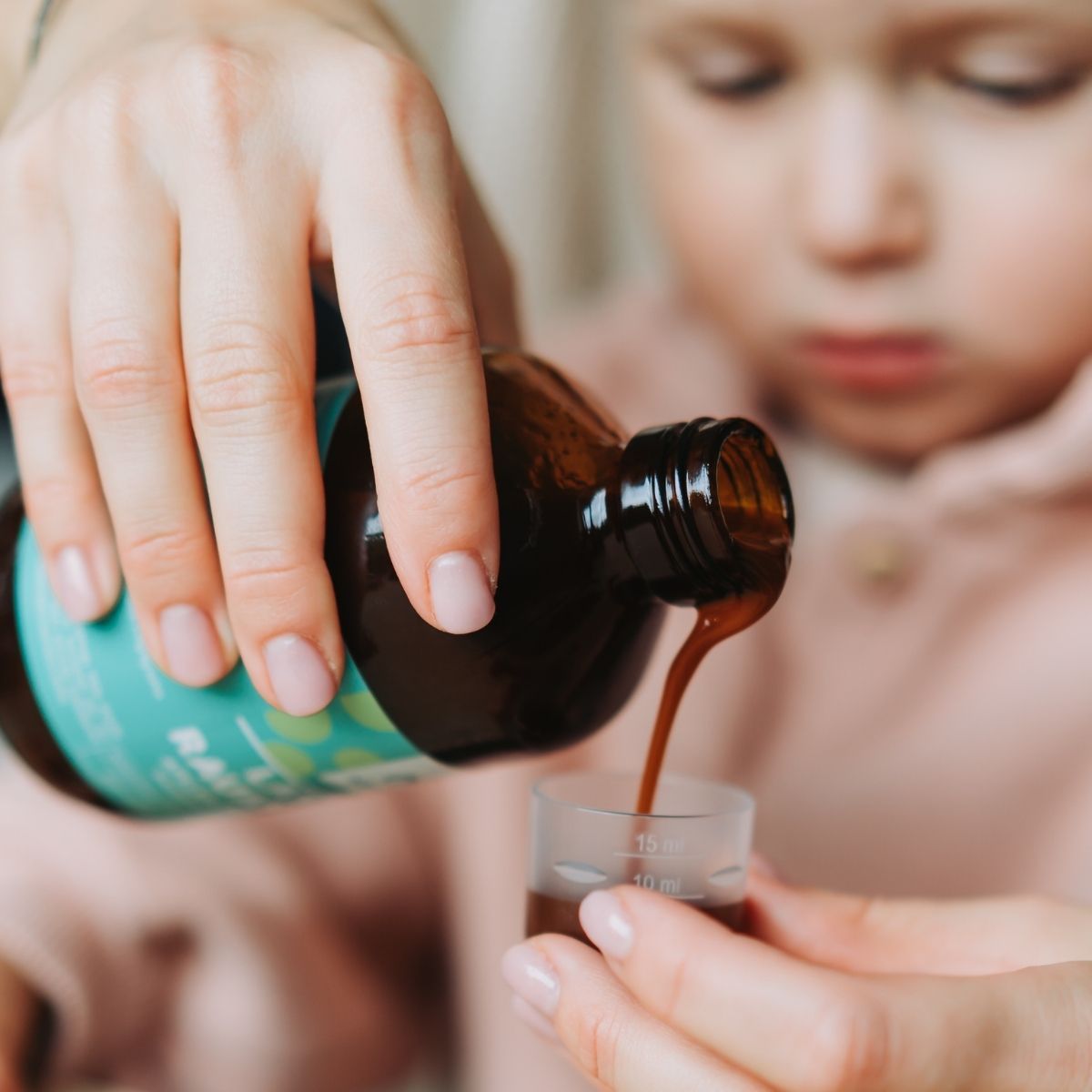 A person pours brown liquid medicine from a bottle into a small clear plastic measuring cup, with a child in the background watching closely as the remedy is prepared. The medicine bottle is dark amber plastic with a white cap and features a simple printed label; text is visible but not readable. Indoor setting, medium shot shows hands and upper torsos.