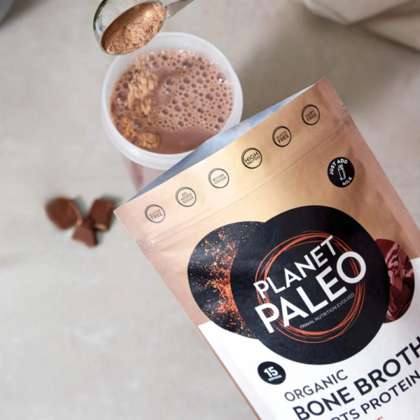 Planet Paleo Chocolate -pussi yläviistosta, edessä vaahtoava suklaajuoma lasissa ja jauhetta valumassa mittalusikasta