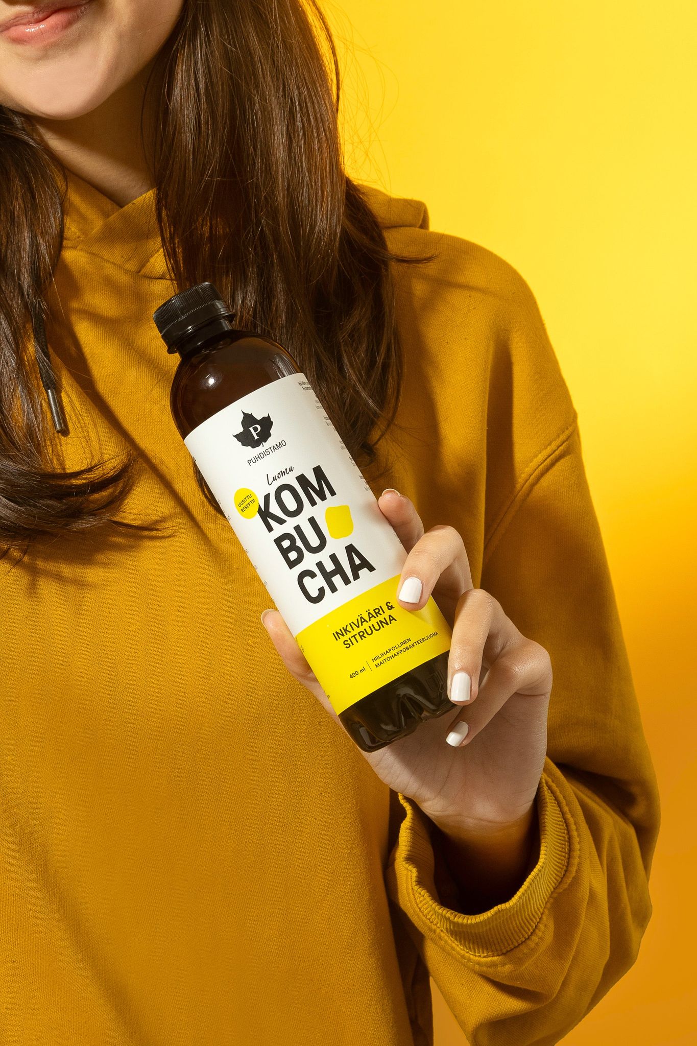 A woman in a yellow sweatshirt holds a clear glass bottle containing kombucha. The bottle has a cylindrical shape and features a yellow and white label with bold, modern typography; brief legible text includes the product name. The label finish is matte. Background is solid bright yellow with evenly lit lighting.