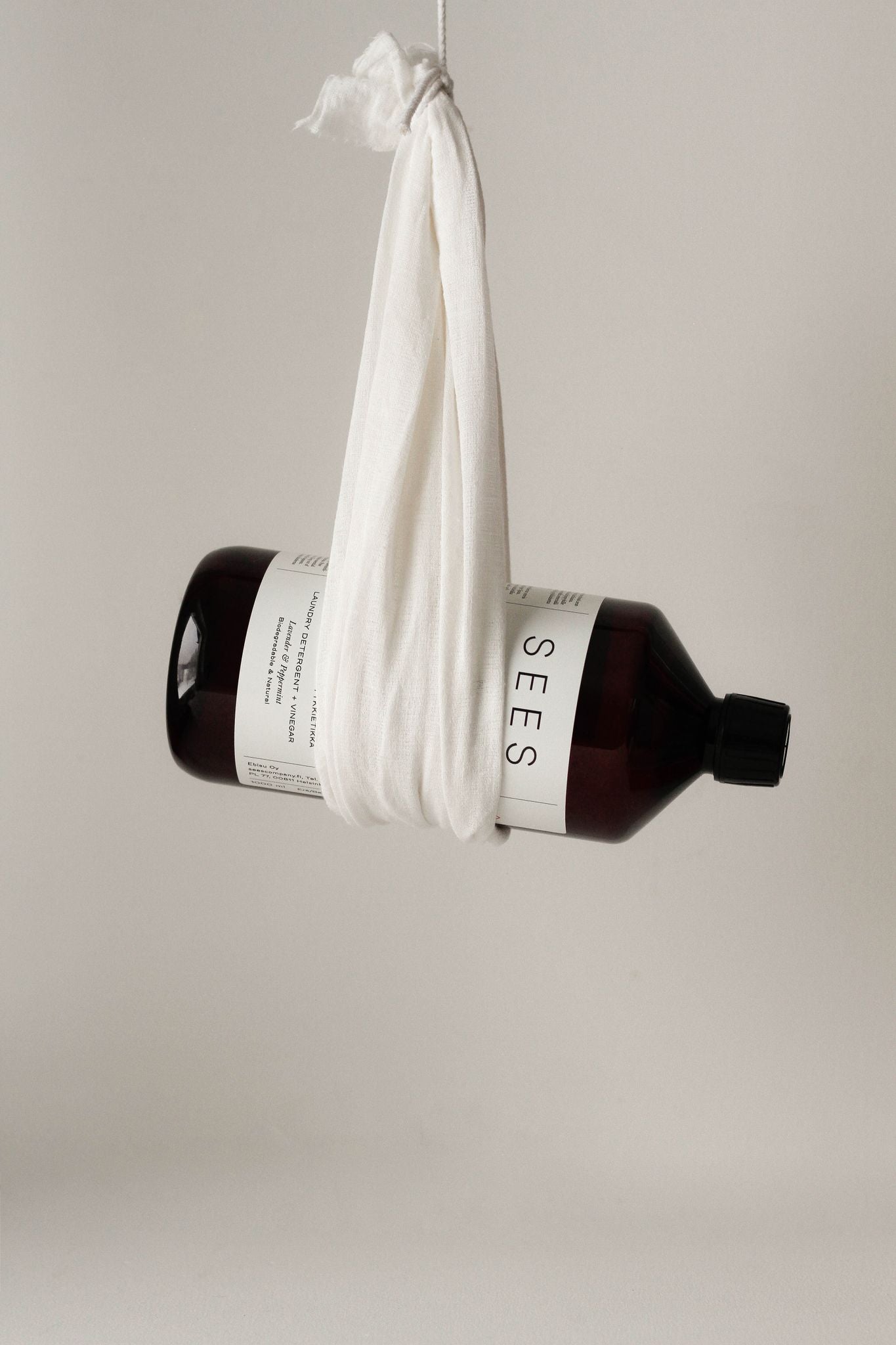 A brown, cylindrical bottle with a matte finish and the word SEES on its label is held horizontally in midair by a white cloth tied in a knot around it. The label uses bold, sans-serif uppercase lettering. There are no people or hands present. The background is plain and light-toned. A brown bottle labeled SEES is suspended horizontally in midair by a white cloth knotted around it, set against a plain light background.