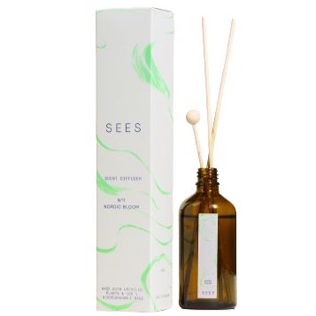 A brown glass bottle with reed diffuser sticks stands beside a white and green SEES box featuring the Nordic Bloom label. The diffuser bottle is cylindrical with a matte finish and several natural-colored wooden sticks inserted. The SEES box has block sans-serif lettering, color blocking in white and forest green, and sharp edges. Both items are positioned on a plain light surface; background is off-white. No people or hands are present in the image.