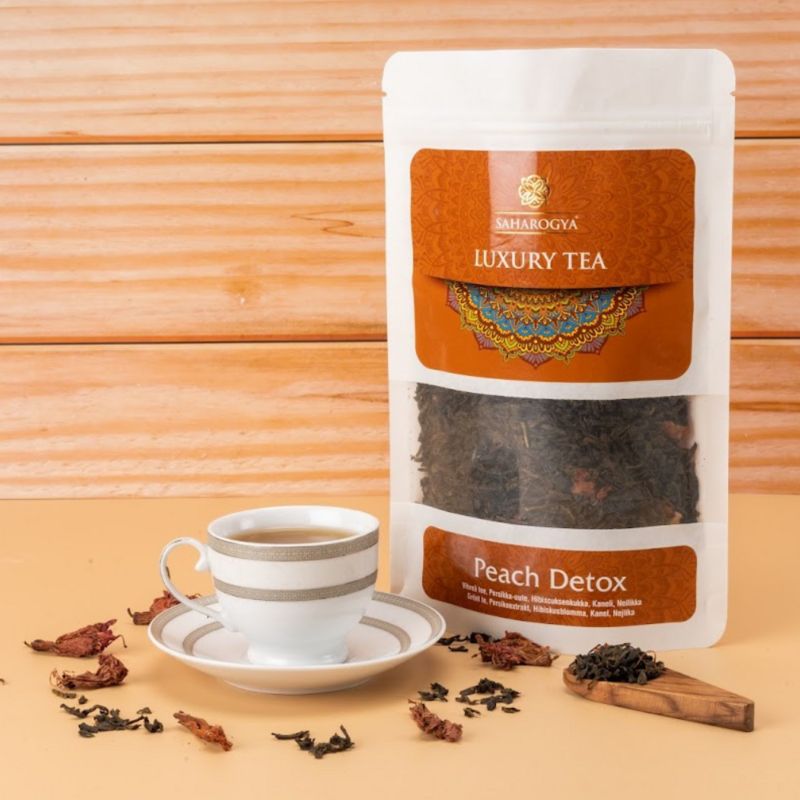 A cup of tea on a white saucer is placed next to a rectangular paper package labeled “Saharogya Luxury Tea Peach Detox” in printed bold serif and script fonts. Loose tea leaves and dried petals are spread on the light wood surface. The background is also light wood. No people or hands are present. Medium product shot, straight-on angle.
