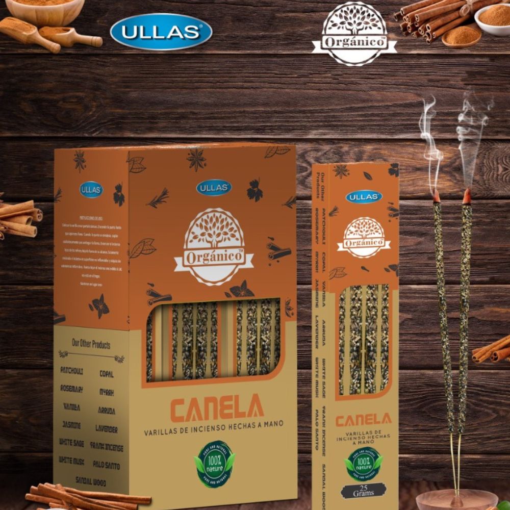 Two boxes of Ullas Orgánico canela (cinnamon) incense sticks are displayed vertically, with two incense sticks burning beside them. Rectangular cardboard boxes feature a matte finish and a brown color palette; packaging includes printed images of cinnamon sticks and white sans-serif label text with green elements. Cinnamon sticks and brand logos enhance the natural atmosphere in the background. No people or hands visible. The setting is a flat tabletop surface photographed from above at a slight angle.