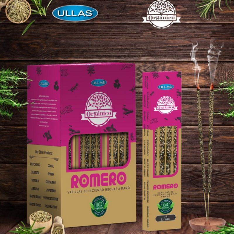 Boxes and sticks of Ullas Orgánico Romero incense are displayed on a wooden surface, with two incense sticks burning and light smoke rising. The packaging is rectangular in shape, colored pink and beige with a matte finish. Botanical illustrations appear on the boxes. Sans-serif text lists organic ingredients and product information in clear lettering. No people or hands present. Medium shot captures all items on the wood background.