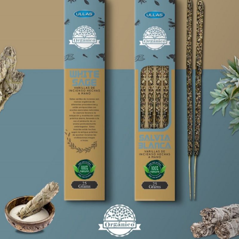Two boxes of Ullas Orgánico white sage incense sticks, each displaying the fragrant sticks inside. Boxes are rectangular with a matte finish and have images of sage on the packaging. Sage leaves, a bundled sage stick, and a round bowl with light sand are arranged nearby. The Orgánico logo is visible on both boxes. The background is blue and tan. No people or hands are present in the image.