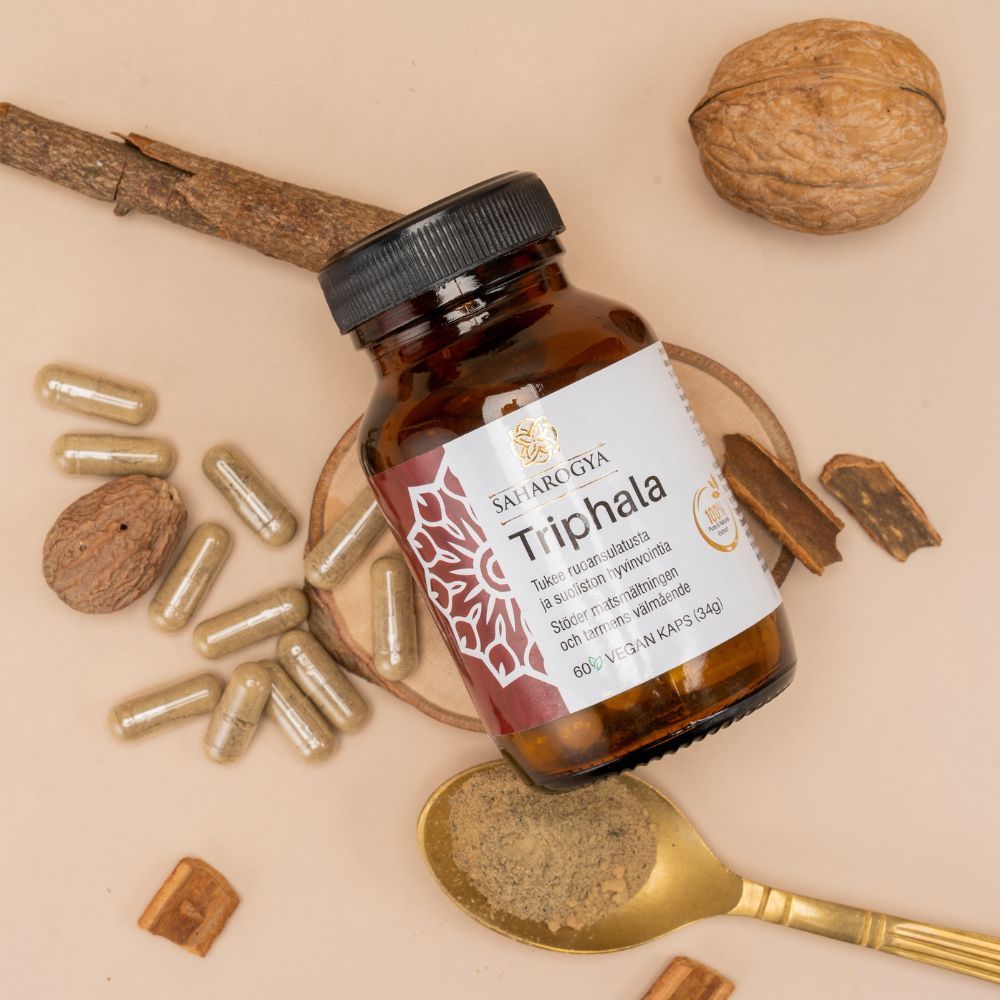 Saharogya Triphala 500 mg; pullo makaa alustalla kapseleiden, kultaisen jauhetäytteisen lusikan, saksanpähkinän, kanelitankojen, muskottipähkinän ja muiden ayurvedisten ainesosien ympäröimänä; lieriön muotoinen, läpinäkymätön ja mattapintainen etiketti lihavoiduin versaalikirjaimin "Triphala"; laakakuva neutraalilla taustalla, ei ihmisiä tai käsiä.