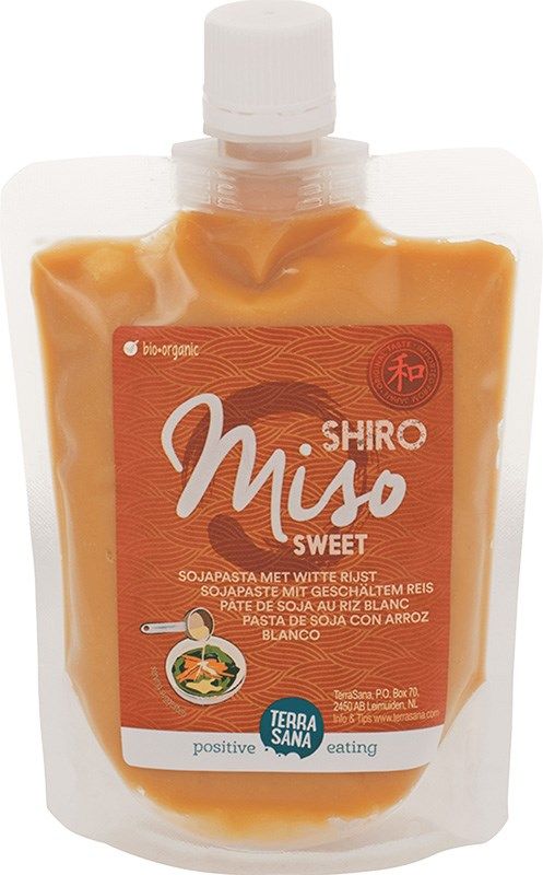 A stand-up pouch of TerraSana Shiro Miso Sweet paste with an orange matte label, white screw cap at the top, clear plastic packaging showing beige miso paste inside. The front label displays legible product name and ingredients in multiple languages, a simple illustrated bowl of soup, and block sans-serif typography. Product is photographed against a plain background with no people visible.