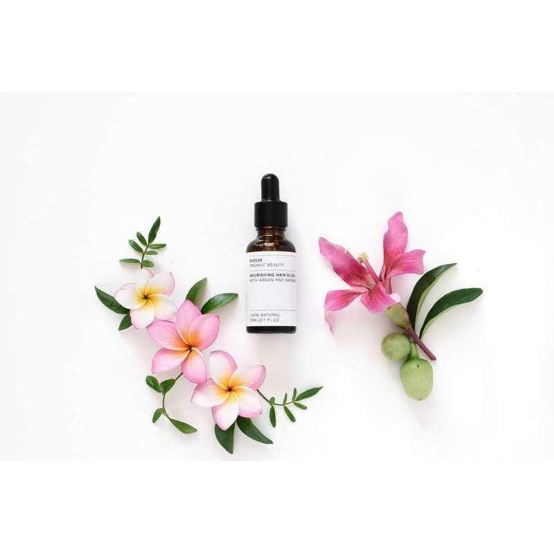 Evolve Nourishing Hair Elixir Kosteuttava Hiusseerumi -tippapullo valkoisella pinnalla, ympärillä värikkäitä vaaleanpunaisia ja valkoisia frangipanikukkia, tuuheita vihreitä lehtiä sekä hento vaaleanpunainen liljankukka; sylinterimäinen, kirkas ja kiiltäväpintainen pullo hopeanvärisellä korkilla ja lasisella tippaputkella; ei luettavaa etikettitekstiä tai brändäystä näkyvissä; ei ihmisiä tai käsiä kuvassa.