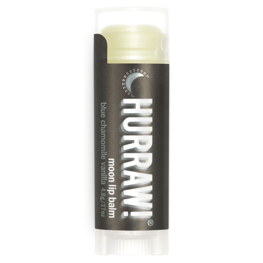 Hurraw! Huulivoide Moon Balm -tuubi, tummansävyinen, sylinterinmuotoinen, mattapinta; lihavoitu valkoinen groteskiteksti "Hurraw! moon lip balm blue chamomile vanilla", vaaleaa voidetta näkyvissä reunan yläpuolella