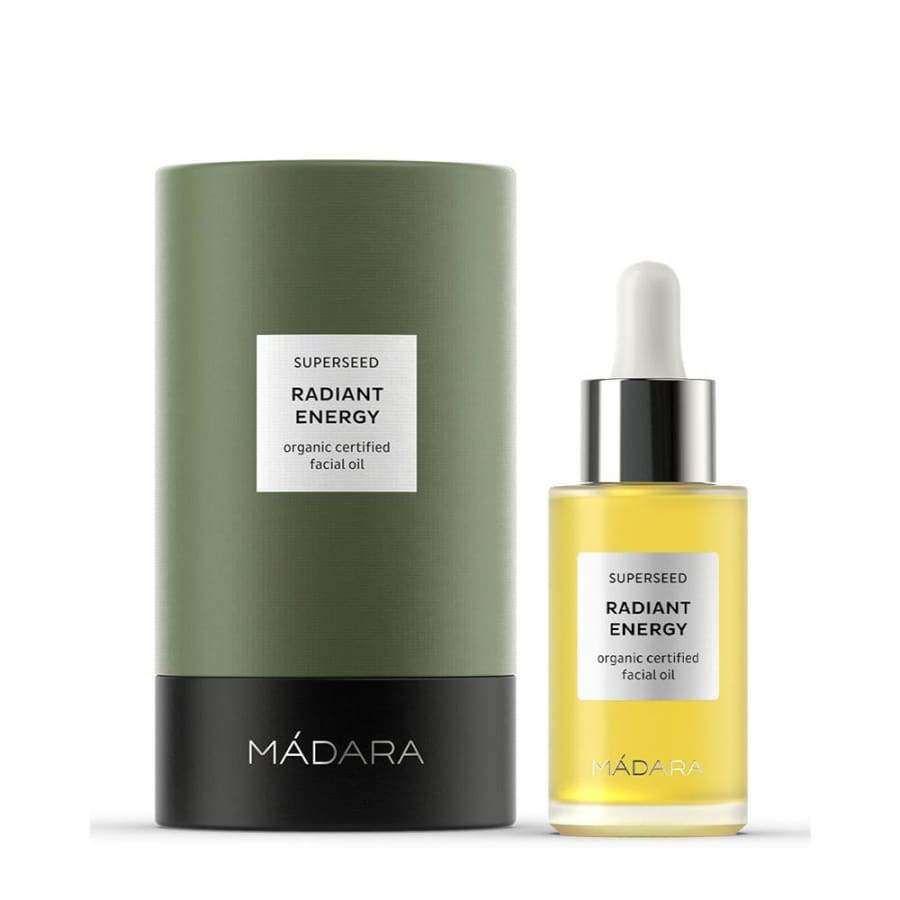 MÁDARA Radiant Energy facial oil in a clear glass bottle with a white dropper cap, placed beside a tall, green and black cylindrical box. The label on the bottle features sans-serif text stating SUPERSSEED Radiant Energy Organic Certified Facial Oil. The background is neutral. No people or actions are present in the image.