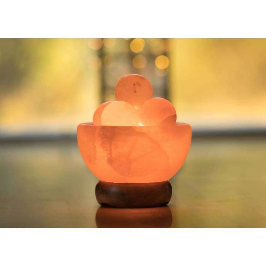 A glowing Himalayan salt lamp shaped like a rounded bowl holds several polished salt stones. The bowl and stones are pink-orange with a matte, crystalline surface. The lamp sits on a circular wood base with a smooth texture. No labels or text are visible. Background is blurred in warm yellow tones, no discernible setting, and the shot is close-up.