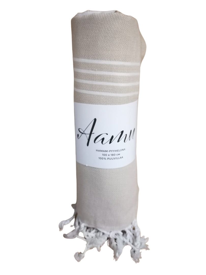 Beige rolled-up Aamu towel with white stripes and fringed ends, labeled on a white wrapper. The wrapper displays the “Aamu” name in a simple sans-serif font. The towel is cylindrical in shape, matte textured fabric, no visible branding on the towel itself. Photo taken against a plain white background, no people or props present.