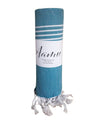 A teal rolled-up towel with white fringe and stripes at one end, featuring a white Aamu label wrapped around it. The material looks to be fabric with a matte finish. The label displays “Aamu” in simple sans-serif lettering. No people or hands are present in the shot. The background is plain and neutral, emphasizing the product.