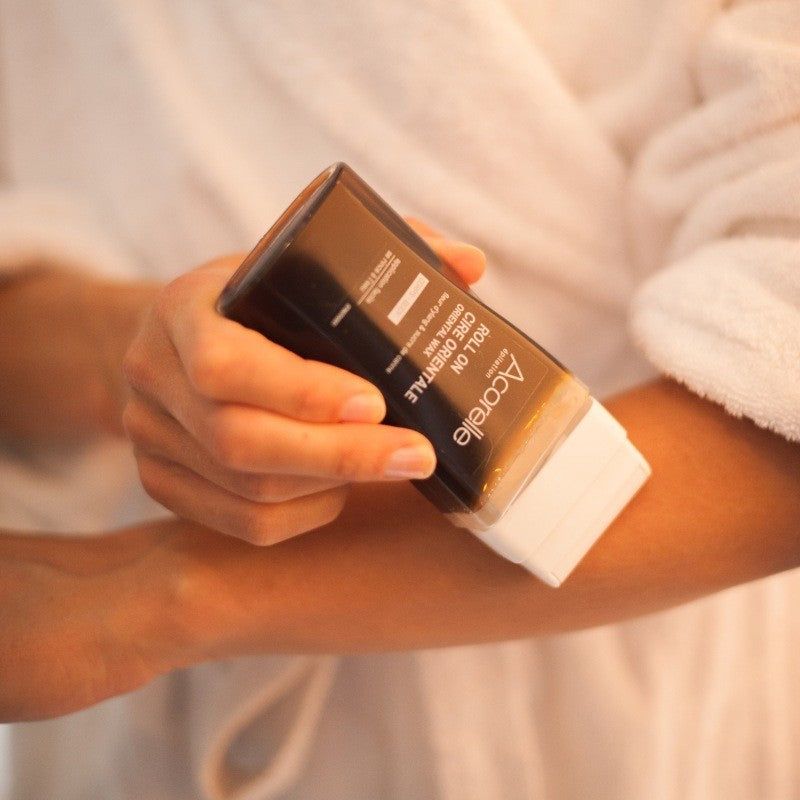 Acorelle Sokerointivaha Roll-On: valkoiseen kylpytakkiin pukeutunut henkilö levittää roll-on-deodoranttia kyynärvarteensa pitäen pulloa ylösalaisin tasaisen levityksen varmistamiseksi; tuote on sylinterimäinen roll-on-deodoranttipullo valkoisella korkilla ja puoliläpinäkyvällä rungolla; etiketissä lihavoitu musta teksti "Acadia"; kuva on otettu neutraalissa sisätilassa läheltä, keskittyen käteen ja tuotteeseen.