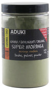 A clear plastic jar labeled ADUKI Super Moringa organic powder, 140g. The jar has a cylindrical shape and contains fine green powder. The black and yellow label includes block lettering, multilingual text, and a red note: Grown by organic family farm. Label placement is centered on the jar body. Background is plain white with even lighting.