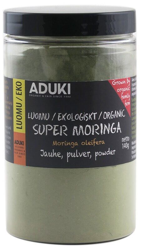 A clear plastic jar labeled ADUKI Super Moringa organic powder, 140g. The jar has a cylindrical shape and contains fine green powder. The black and yellow label includes block lettering, multilingual text, and a red note: Grown by organic family farm. Label placement is centered on the jar body. Background is plain white with even lighting.