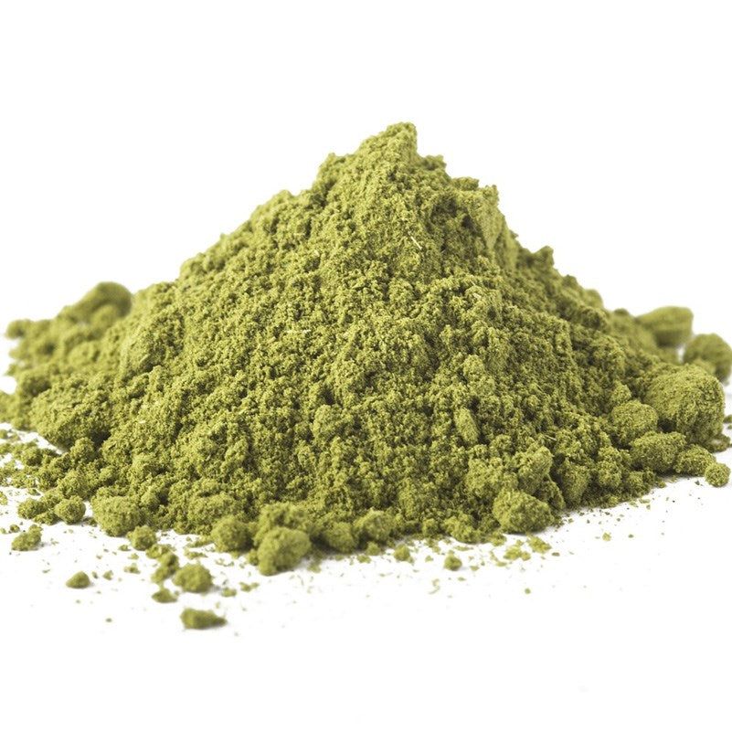 A small mound of vibrant matcha powder is spread on a white surface, with some loose granules scattered around the main pile. The product is finely ground green powder with a matte texture and bright green color. There is no visible packaging or labeling, no typography present, and no people or hands in the image. The photo uses a neutral background for contrast and appears to be a close-up shot taken from above.