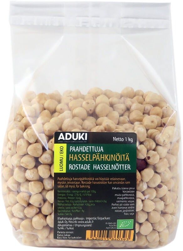 A clear plastic bag filled with roasted hazelnuts. Rectangular, transparent packaging with a glossy finish. The label is black and white with bold sans-serif “ADUKI” text; other visible details include Finnish and Swedish wording, a green eco symbol, and net weight of 1 kg. Neutral background; no people or hands present.