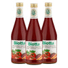 Three bottles of Biotta Organic Vegetable Cocktail juice with red liquid inside, each bottle featuring images of tomatoes and vegetables on the front label. Bottles are made of clear glass with a glossy finish, tapered necks, and metal caps. Labels show white background and bold green lettering for the brand name. White surface background; no people or hands visible; standard product shot.