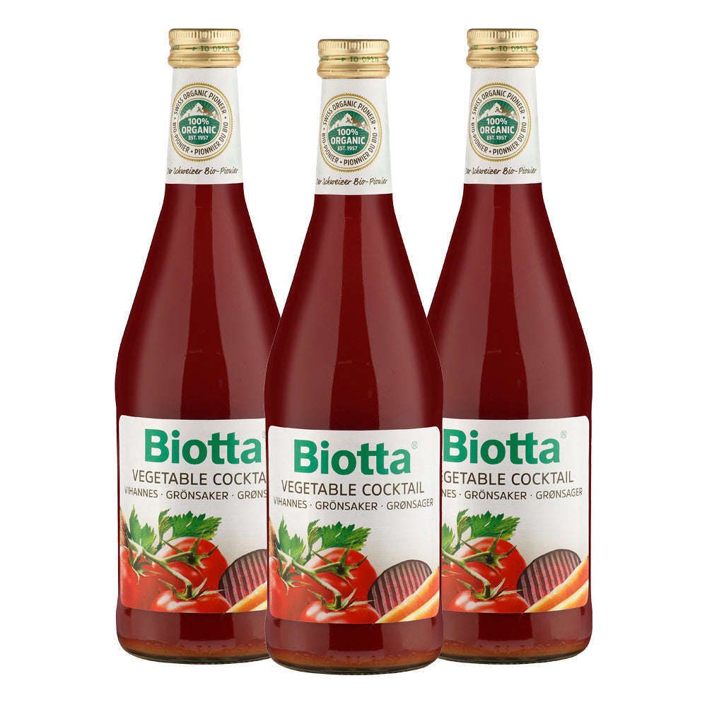 Three bottles of Biotta Organic Vegetable Cocktail juice with red liquid inside, each bottle featuring images of tomatoes and vegetables on the front label. Bottles are made of clear glass with a glossy finish, tapered necks, and metal caps. Labels show white background and bold green lettering for the brand name. White surface background; no people or hands visible; standard product shot.