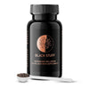 A black, cylindrical supplement bottle with a matte surface finish displays the label “Black Stuff,” a tree graphic, and text including “Microbiome Wellbeing Fulvic Acid Rich Supplement” in clear sans-serif fonts. It stands beside a glossy white spoon containing coarse, dark granules. The objects are arranged on a plain, light background. No people or hands visible.