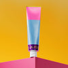 A colorful tube of cream featuring pink, blue, and metallic purple sections stands upright on a bright pink corner against a yellow background. The tube is made of glossy plastic, cylindrical with a flat flip-top cap at the base. Packaging displays segmented colors without gradients. No visible people or hands. Brand label text uses bold sans-serif font; only product name and logo are legible on the front.