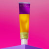 A colorful tube labeled The Juice Moisturizing Power Fluid stands upright on a shiny, solid purple surface. The tube features a simple cylindrical shape and has clear, bold lettering on the label. The background displays a bright pink gradient fading from top to bottom. No people or hands are visible in the image.