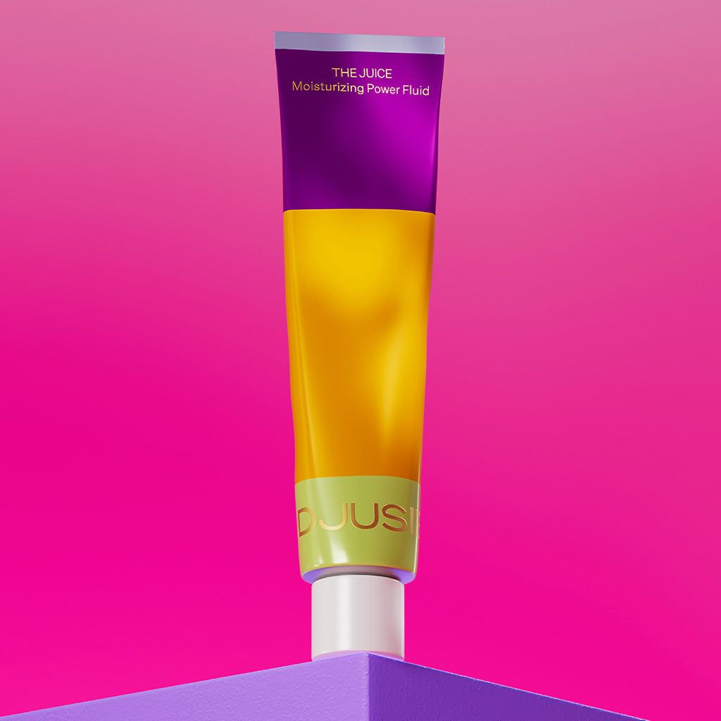 A colorful tube labeled The Juice Moisturizing Power Fluid stands upright on a shiny, solid purple surface. The tube features a simple cylindrical shape and has clear, bold lettering on the label. The background displays a bright pink gradient fading from top to bottom. No people or hands are visible in the image.