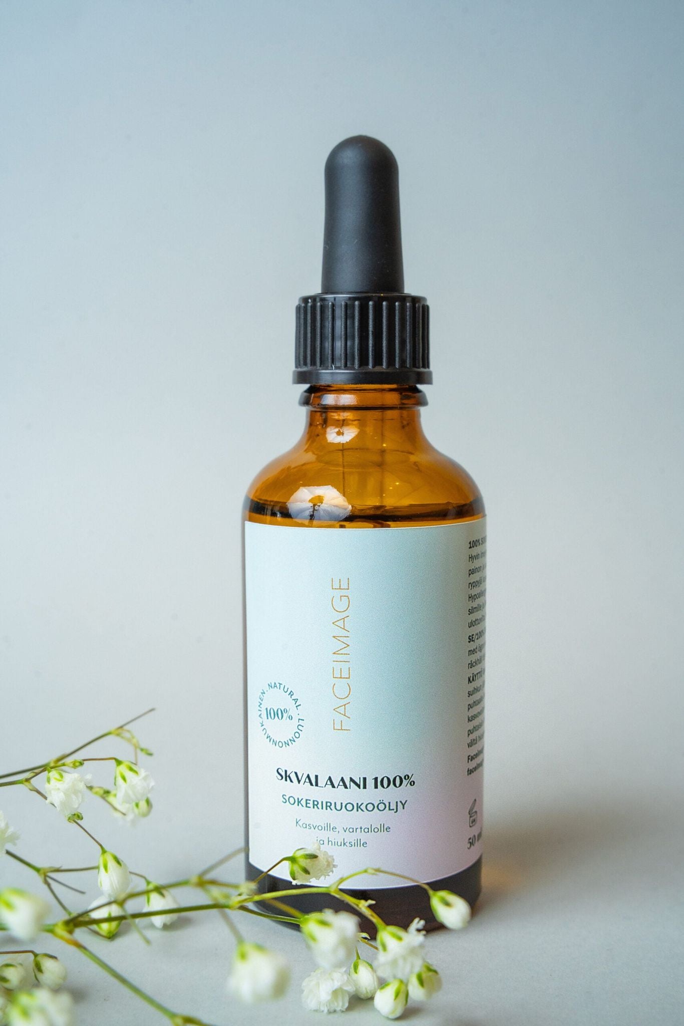 A brown glass dropper bottle with a white cap is positioned upright. The label features the text “Squalane 100%” in clear, sans-serif font. White flowers are arranged in front of the bottle. The surface beneath is light-colored and smooth, and the plain background is light blue. No people or hands are present in the image.
