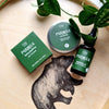 Three green-labeled Fiini Perkele mens grooming products rest on a wooden tray featuring a bear illustration, with a leafy plant partially visible at the top. The products have matte black or dark containers: one pump bottle, one dropper bottle, and one jar. White sans-serif text reads “Fiini Perkele” on each label. Overhead shot; no people in frame.