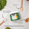 A jar labeled Green Superfood is surrounded by wooden spoons holding vibrant powders, with text highlighting ingredients such as Bacillus Coagulans bc30 probiotic, Supermaca, and Lion’s Mane. The container is cylindrical with a matte finish and features clear block lettering. The background includes whole and sliced fresh greens and fruits on a flat surface. No people or hands are present in the shot.