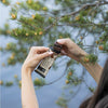 A person opens a chocolate protein bar outdoors. The hands are visible holding the bar, which has a rectangular shape and is partially wrapped in glossy packaging with brown coloring. Simple sans-serif label text is partially legible on the wrapper. A pine tree trunk and still water appear in the background.