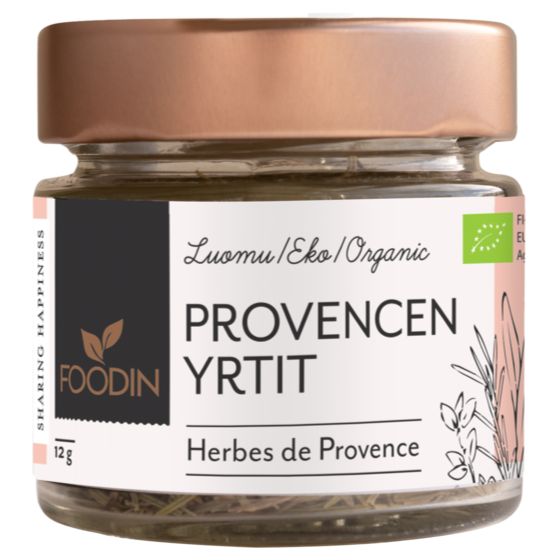 A glass jar with a rose gold metallic screw-top lid labeled FOODIN PROVENCEN YRTIT and Herbes de Provence features a Luomu/Eko/Organic label and charming herb illustrations. The transparent cylindrical jar contains a dried herb mix. The paper label is matte with sans-serif type, additional small text, and stylized botanical drawings. Plain white background, no people visible.