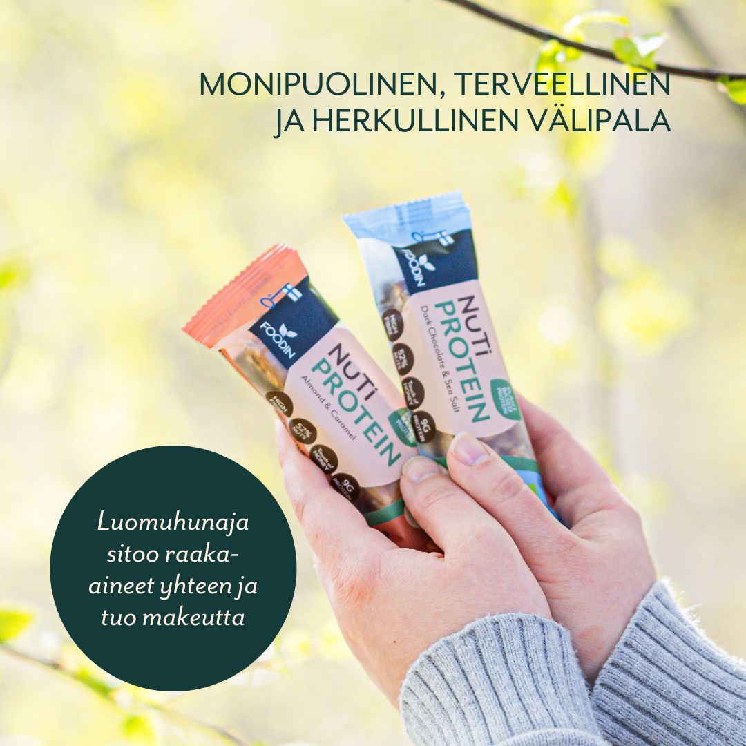 Foodin NUTI Luomu Pähkinäpatukka Protein, Almond & Caramel; kaksi kättä pitelee kahta pähkinäproteiinipatukkaa ulkona epätarkkaa vihreää taustaa vasten; patukat mattapintaisessa pakkauksessa, kumpikin suorakulmainen ja foliokäärintäinen; pakkauksessa lohkomaista suomenkielistä tekstiä; pääetiketti esittää tuotenimen ja mainitsee luomuhunajan, pähkinät sekä välipalaominaisuudet; ei muita esineitä tai ihmisiä kuvassa.
