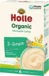 Box of Holle Organic Wholegrain 3-Grain Cereal with a cartoon frog illustration, yellow corn, and wheat graphic on matte cardboard packaging. Rectangular box shape. Clear bold sans-serif product name; icons indicate dairy-free, gluten-free, 6+ months suitability, and climate positive status. No people or hands present. Plain background with no visible setting.