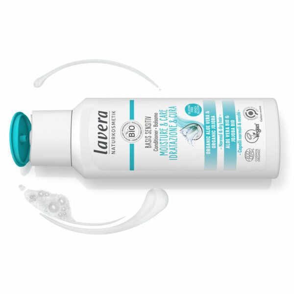 White bottle of Lavera Moisture & Care lotion with turquoise cap, laying on its side. Some lotion is spilled near the bottle. The label highlights vegan product details in English and Italian with blue and black text. Bottle has a cylindrical shape, glossy finish, and printed label with sans-serif typography. Plain background, no people or additional objects visible. Camera angle shows full product from the side in a close-up shot.