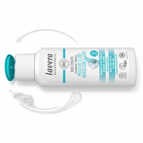 Valkoinen Lavera Moisture & Care -hoitoainepullo turkoosilla korkilla makaa kyljellään, vieressä hieman läikkynyttä hoitoainetta; etiketissä vegaanituotetiedot englanniksi ja italiaksi sinisellä ja mustalla tekstillä; pullo on lieriömäinen kiiltäväpintainen, groteskikirjasin painettuna etikettiin; tausta yksinkertainen, ei ihmisiä tai muita esineitä; kamerakulma näyttää tuotteen kokonaan sivulta läheltä kuvattuna.