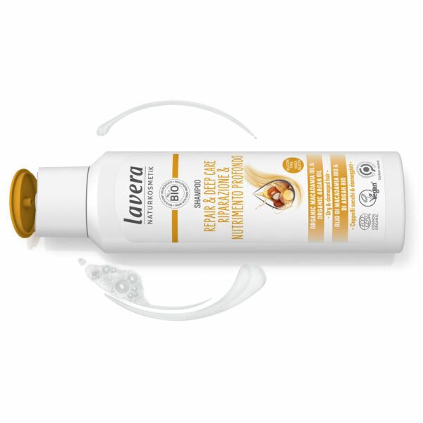 White bottle of Lavera Repair & Deep Care Shampoo with an open yellow cap. The container has a smooth, matte finish and curved edges. Black and gold text is printed on the label, which includes words about argan oil and bio certifications. The bottle is placed on a white background next to two smears of creamy shampoo. No people or hands are visible in the image.