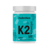 A turquoise plastic bottle with a matte finish and a cylindrical shape; white screw-on cap; label displays “makrobios K2 Vitamiini 100 mikro g” with additional Finnish text in clear sans-serif typography; the label includes abstract shapes. The product is photographed against a plain, evenly lit background with no people visible.