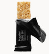 An open black wrapper labeled Maurten Solid 160 reveals a rectangular energy oat bar partially slid out, showing its textured, grainy surface. Wrapper is matte black with white sans-serif text. The oat bar has a light brown color and visible grain texture. No other products or props are present; background is plain and neutral. No people or hands in frame; shot is close-up with focus on the product.