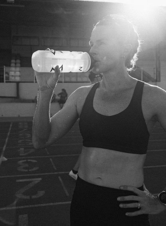 A woman in athletic wear hydrates with a water bottle on a track field, with sunlight shining behind her for a refreshing boost. Plastic water bottle, clear material with visible liquid. Athletic clothing includes leggings and sleeveless top, possibly synthetic fabric. Track surface is red, outdoor setting, natural light shines from upper background. No label text is legible. Medium full shot angle.