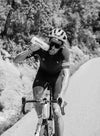 A cyclist wearing a helmet and sunglasses rides uphill on a road, drinking from a water bottle, with trees and bushes in the background. The black and white image shows one person in athletic clothing. Surface details of the bike are visible but not branded. There is no packaging or prominent text in the image. Outdoor setting, action shot, full body visible.