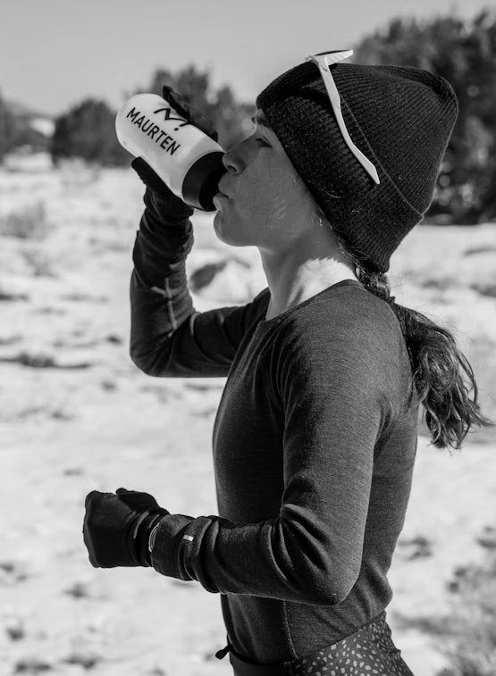 A person in a beanie and gloves drinks from a Maurten water bottle outdoors on a sunny day. The bottle is cylindrical, semi-transparent with a matte finish, and features bold black sans-serif “Maurten” text. Sunglasses rest on the person’s head. The background shows trees and sandy ground under bright sunlight.